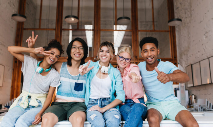 Excited girl in blue shirt showing peace sign enjoying friend's company in good day. Indoor portrait of glad international students fooling around for photo and laughing.