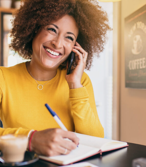 Young afro woman in a coffee shop, smiling and confident, talking on a mobile phone. She is writing in a notebook.